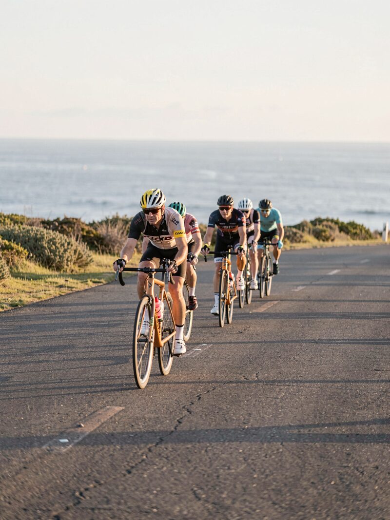 A smooth single-file paceline on a flat coastal road, one rider pulling off the front into the wind, low morning sun