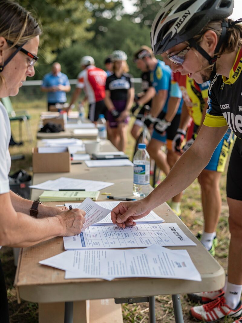 A volunteer behind a folding table checking names on a start list, a short line of riders waiting, wind ruffling the papers